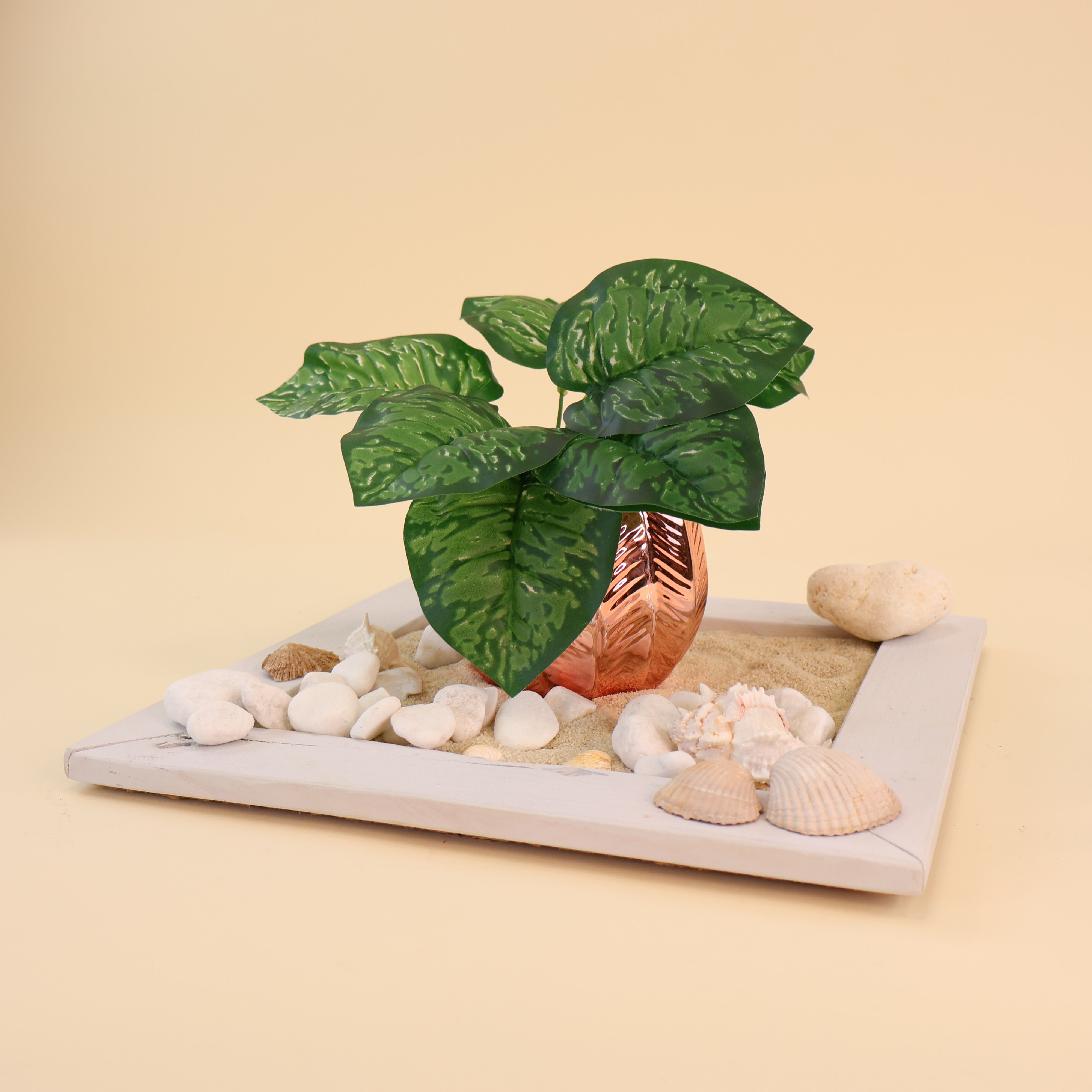 Künstliche Aglaonema mit dunkelgrünen Blättern und hellgrüner Marmorierung dekorativ in einer kupferfarbenen Vase arrangiert. Weiße Kieselsteine, Muscheln und Sand auf einem weißen Tablett verleihen dem Arrangement eine maritime und moderne Optik. Die detailreiche Blattstruktur und die natürliche Farbgebung machen die Kunstpflanze ideal für Vasen, Regaldekorationen und stilvolle Wohnraumbegrünungen.
