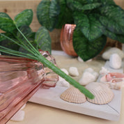 Makroaufnahme des formbaren Stiels einer künstlichen Aglaonema vor einem dekorativen Arrangement mit rosafarbener Glasvase, Muscheln, weißen Kieselsteinen und gemusterten Blättern. Die Nahaufnahme zeigt die hochwertige Verarbeitung und die flexible Gestaltungsmöglichkeit der Kunstpflanze. Die detailreiche Ausführung sorgt für eine natürliche Optik und macht die Blattschmuckpflanze ideal für Vasen, Gestecke und moderne Wohnraumdekorationen.