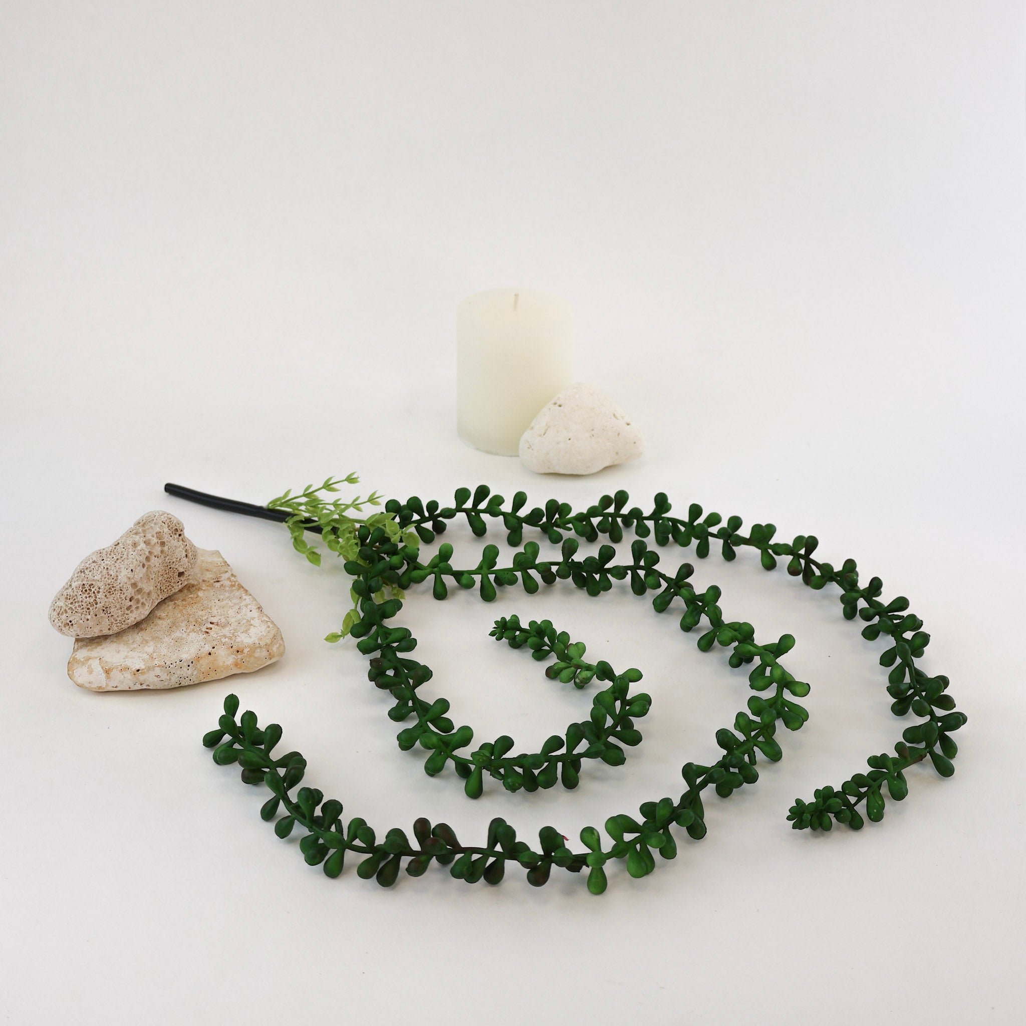 Künstliche Hängepflanze mit langen, geschwungenen Trieben und kleinen dunkelgrünen Blättern auf weißem Hintergrund. Die dekorative Kunstpflanze ist zwischen Natursteinen und einer weißen Kerze arrangiert und eignet sich ideal für Regale, Vasen, Pflanzenbilder und moderne Wohnraumdekorationen.