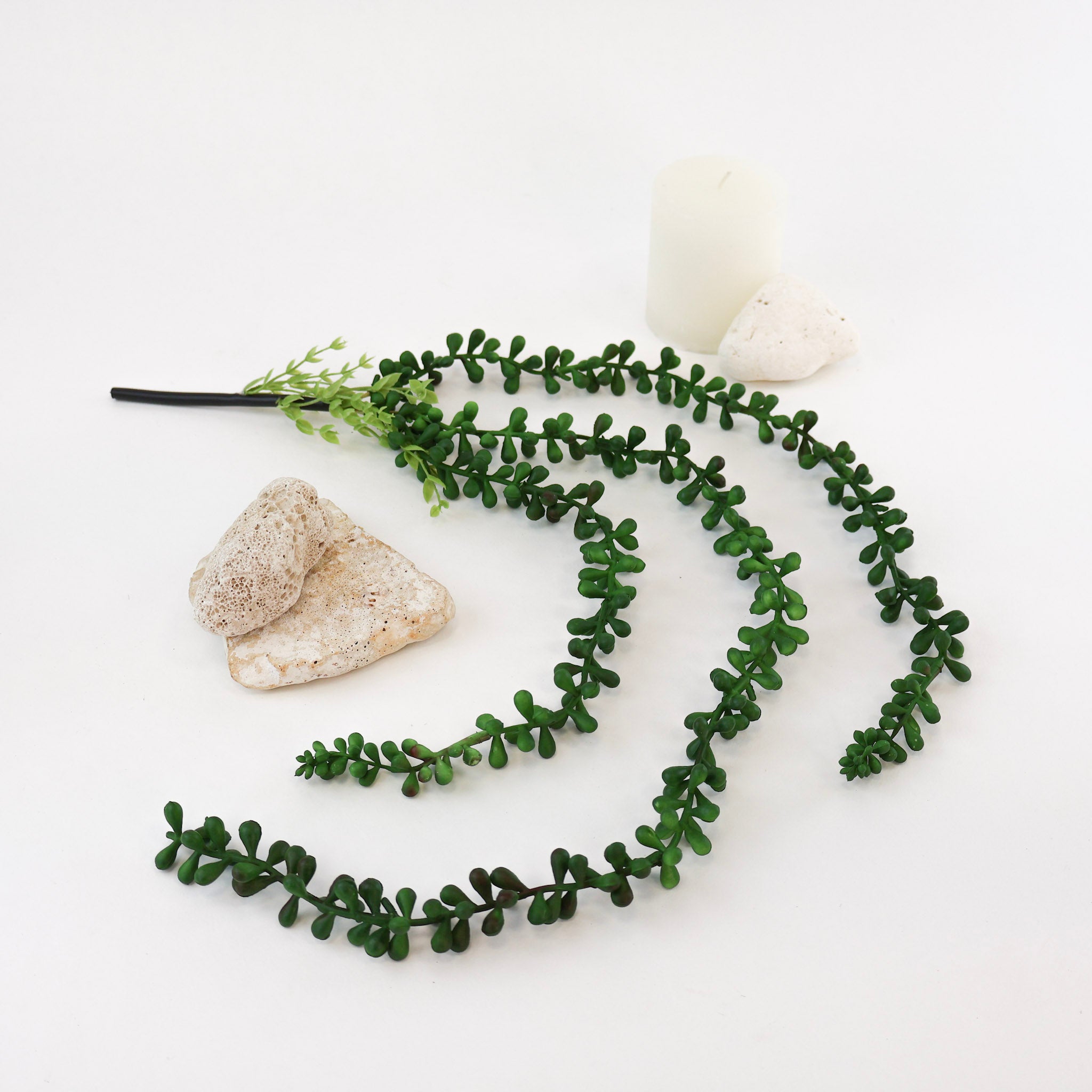 Künstliche Hängepflanze mit langen, elegant herabhängenden Trieben und kleinen dunkelgrünen Blättern auf weißem Hintergrund. Die naturgetreu gestaltete Kunstpflanze ist dekorativ mit hellen Natursteinen und einer weißen Kerze arrangiert und eignet sich ideal für Regale, Vasen, Pflanzenbilder und moderne Wohnraumdekorationen.