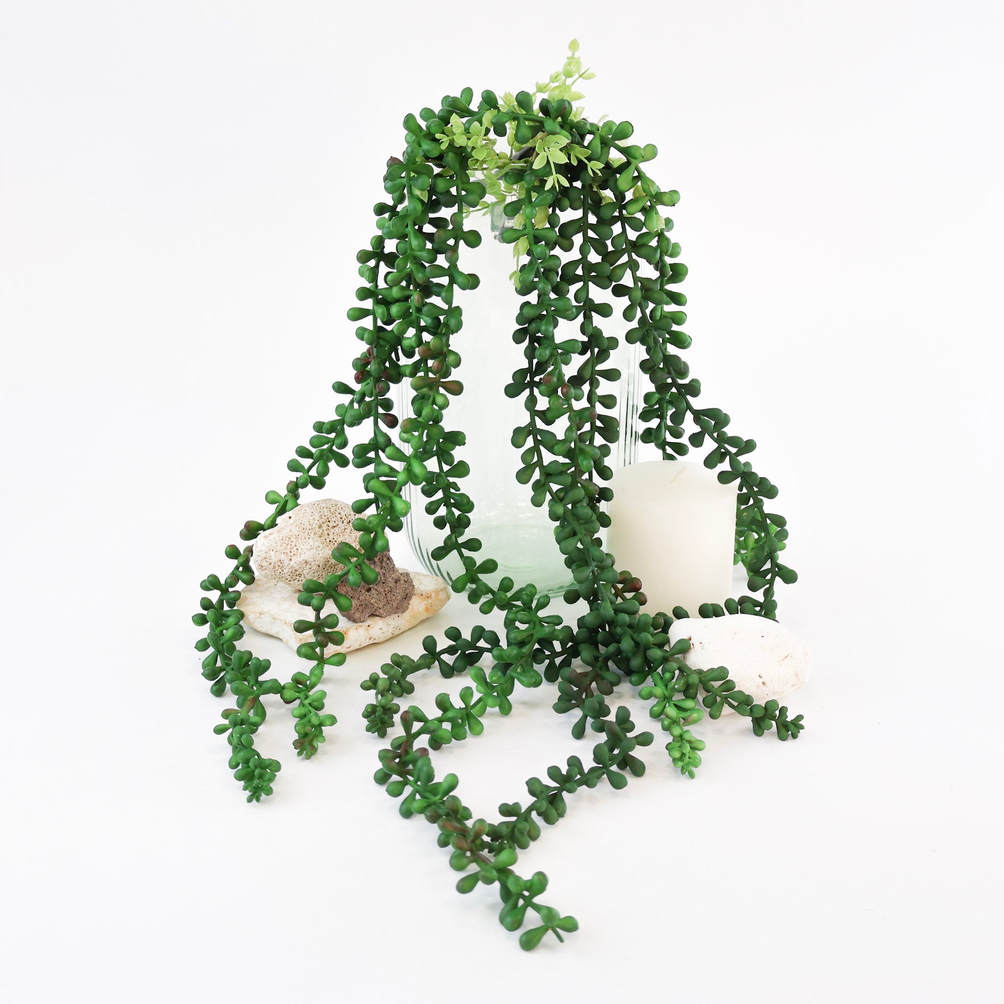 Künstliche Hängepflanze mit langen, herabhängenden Trieben und zahlreichen kleinen dunkelgrünen Blättern in einer dekorativen Glasvase. Die üppige Kunstpflanze fällt elegant über die Vase hinweg und wird von Natursteinen und einer weißen Kerze stilvoll ergänzt. Ideal für Regale, Sideboards und moderne Wohnraumdekorationen.