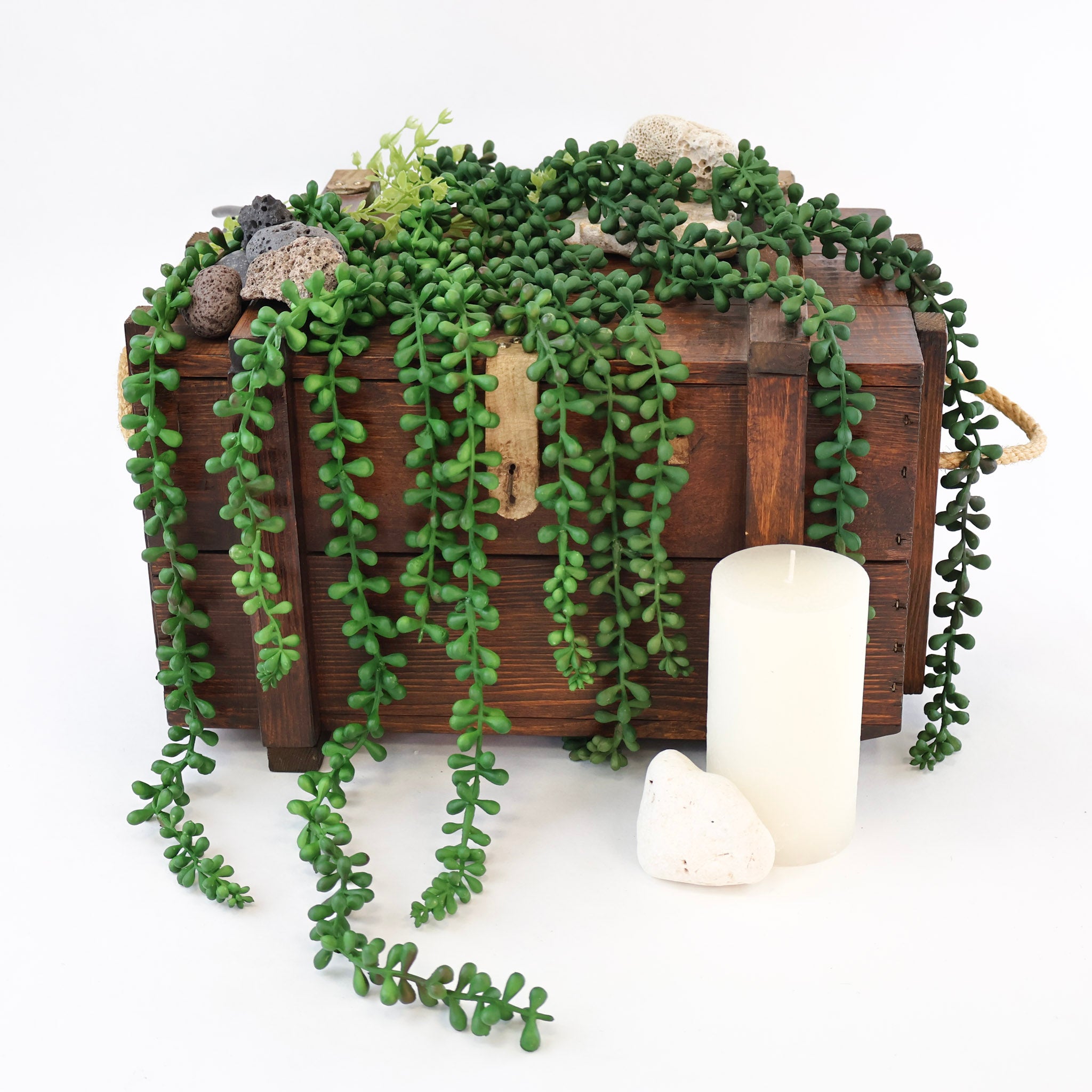 Künstliche Hängepflanze mit langen, dicht bewachsenen Trieben und kleinen dunkelgrünen Blättern dekorativ über eine rustikale Holzkiste drapiert. Weiße Natursteine und eine Kerze ergänzen das Arrangement und unterstreichen die natürliche Optik der herabhängenden Kunstpflanze für Regale, Sideboards und moderne Wohnraumdekorationen.