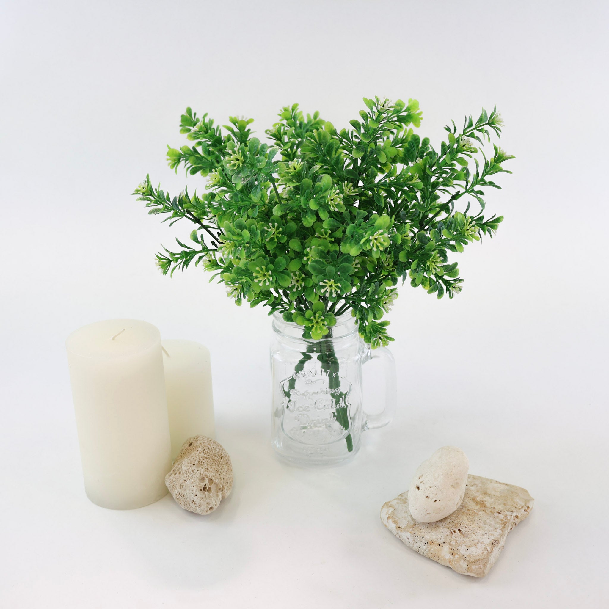 Künstlicher Grünzweig mit kleinen grünen Blättern und feinen weißen Blüten in einem Glasgefäß dekorativ arrangiert mit weißen Kerzen und Natursteinen auf weißem Hintergrund.
