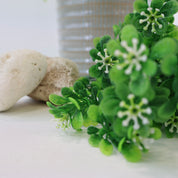 Detailaufnahme eines künstlichen Grünzweigs mit kleinen grünen Blättern und feinen weißen Blüten neben hellen Natursteinen, dekorative Kunstpflanze auf weißem Hintergrund.