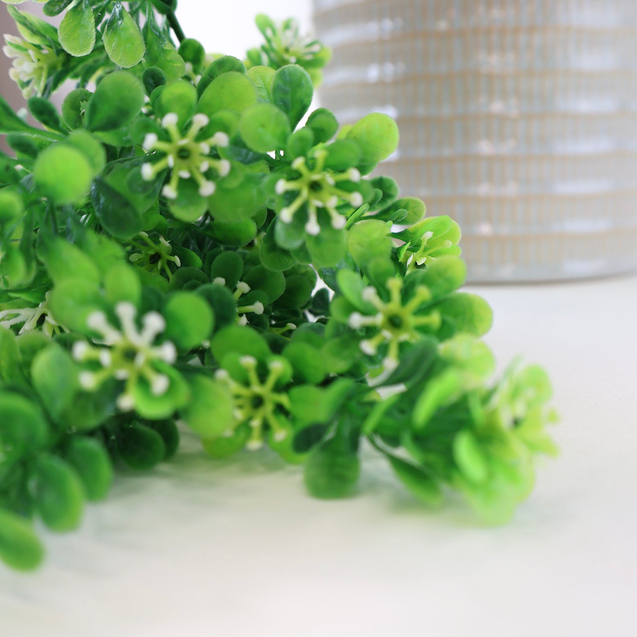 Makroaufnahme eines künstlichen Grünzweigs mit kleinen grünen Blättern und feinen weißen Blüten, dekorative Kunstpflanze mit natürlicher Optik vor hellem Hintergrund.