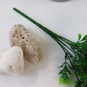 Detailaufnahme künstlicher Grünzweige mit kleinen grünen Blättern und feinen weißen Blüten vor einem strukturierten Keramiktopf und hellen Natursteinen auf weißem Hintergrund.