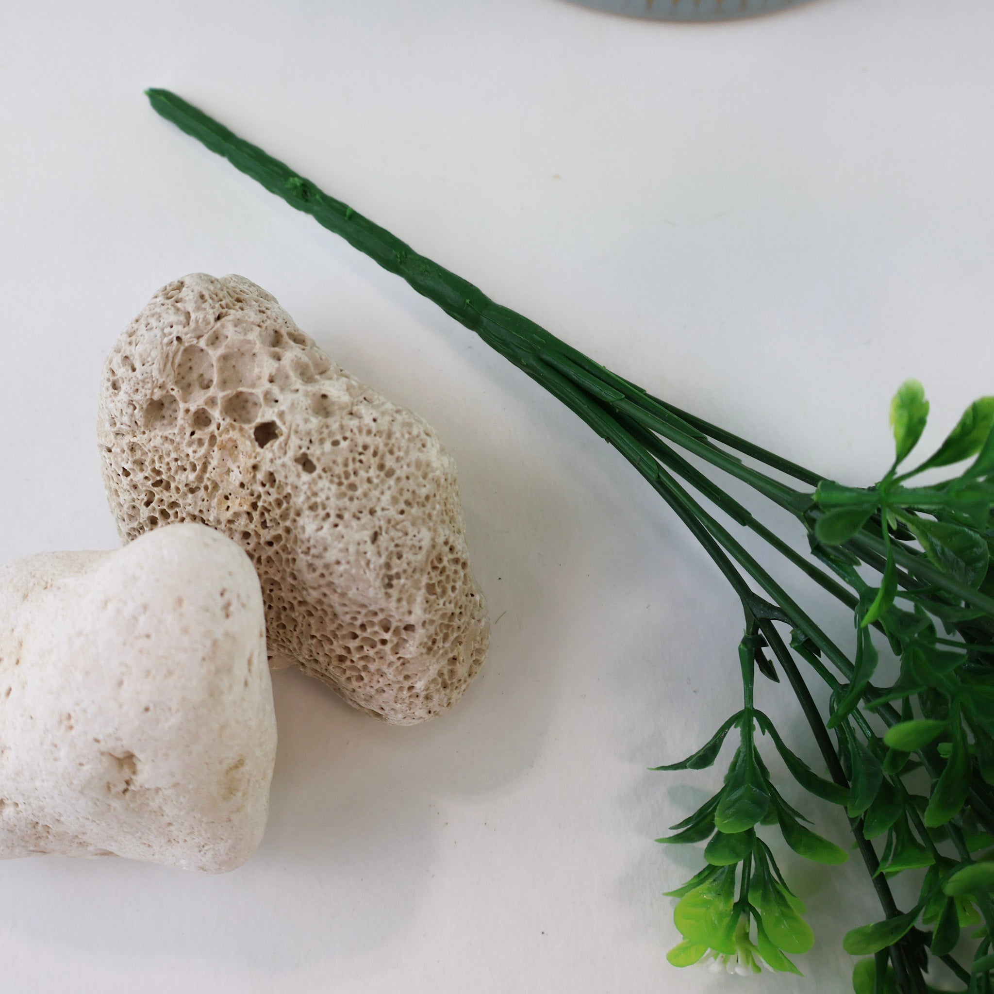 Detailaufnahme künstlicher Grünzweige mit kleinen grünen Blättern und feinen weißen Blüten vor einem strukturierten Keramiktopf und hellen Natursteinen auf weißem Hintergrund.