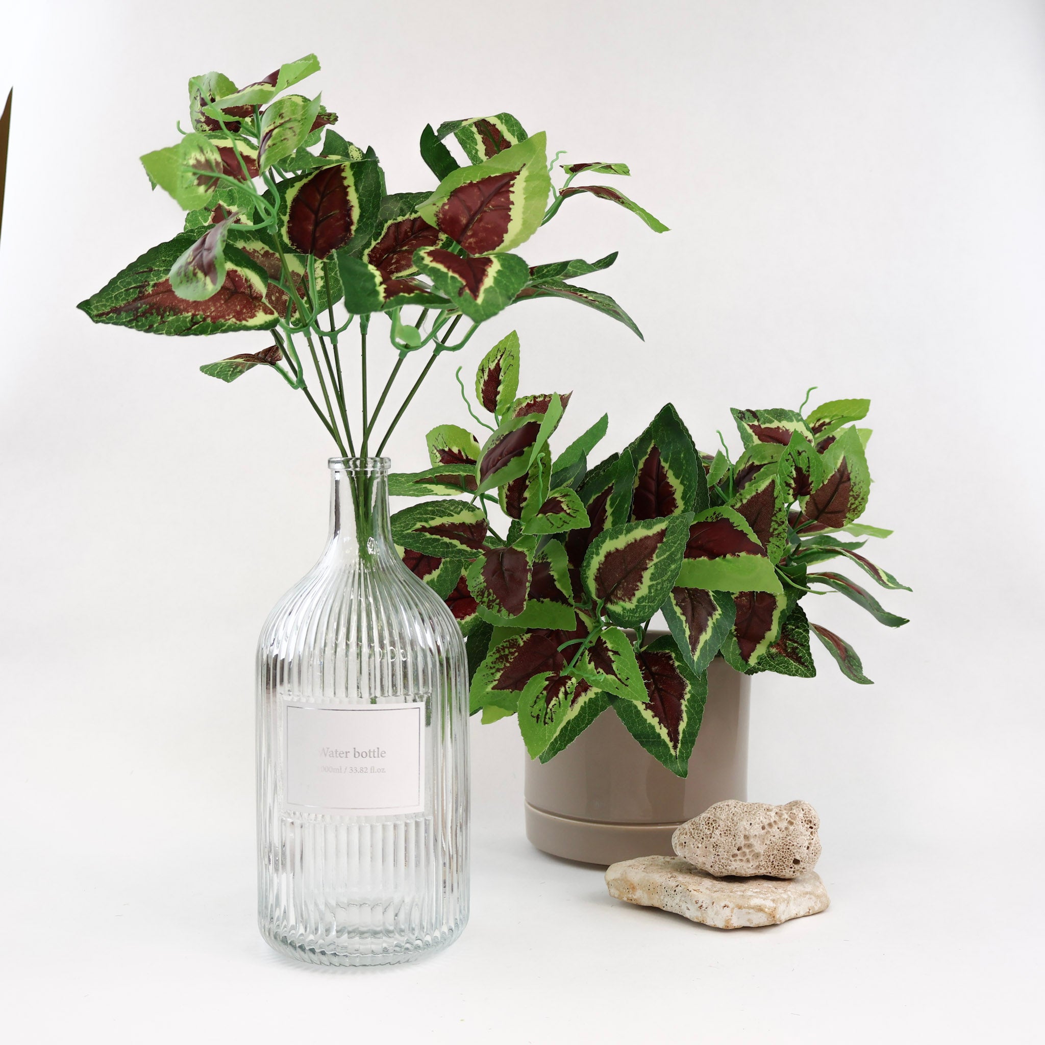 Künstliche Blattzweige mit grün gemusterten Blättern und dunkelroter Blattmitte dekorativ arrangiert in einer Glasflasche und einem Keramiktopf mit Natursteinen auf weißem Hintergrund.