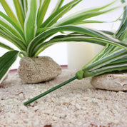 Detailaufnahme künstlicher Graspflanzen mit grün-weiß variierten Blättern auf hellem Dekosand, kombiniert mit Natursteinen und Kerze. Sichtbarer Kunststoffstiel und natürliche Blattstruktur der Kunstpflanze im Fokus.