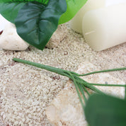 Detailaufnahme des grünen Stiels einer künstlichen Blattpflanze auf hellem Sand mit weißen Kerzen und Natursteinen, dekoratives Arrangement im natürlichen Stil.