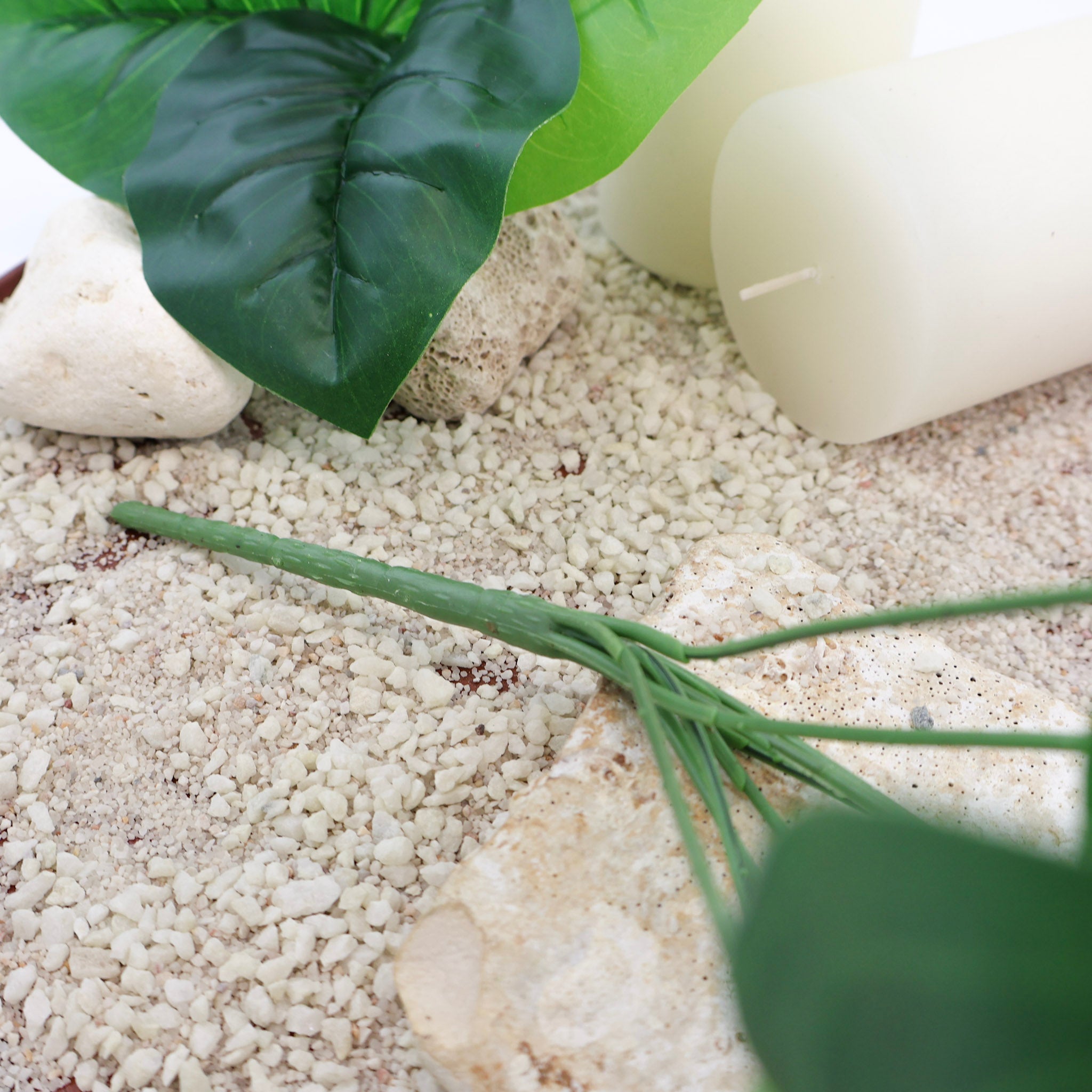 Detailaufnahme des grünen Stiels einer künstlichen Blattpflanze auf hellem Sand mit weißen Kerzen und Natursteinen, dekoratives Arrangement im natürlichen Stil.
