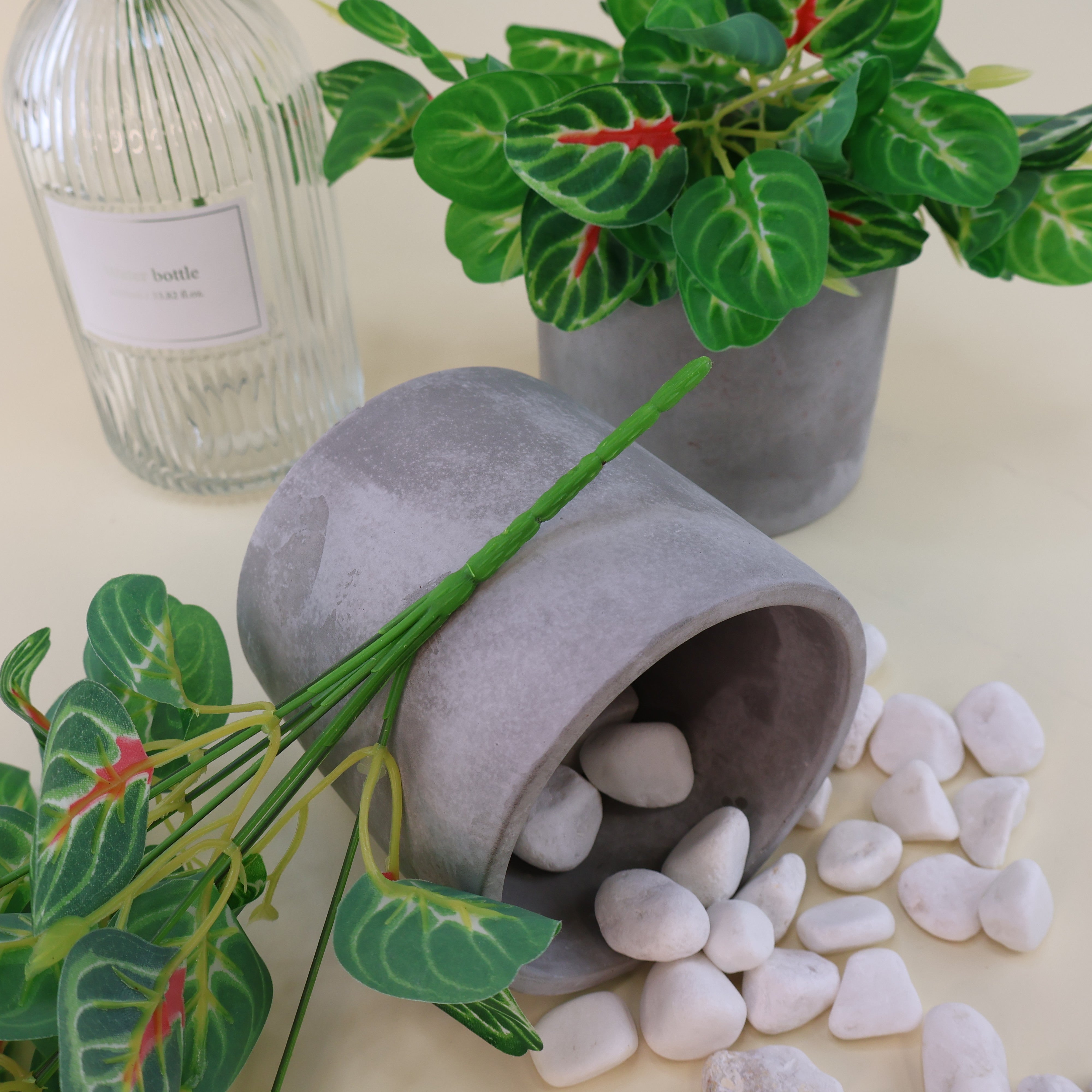 Detailaufnahme einer künstlichen Grünpflanze mit herzförmigen Blättern in verschiedenen Grüntönen und roten Blattakzenten. Die Kunstpflanze ist mit einem grauen Keramiktopf, weißen Dekosteinen und einer strukturierten Glasvase dekorativ arrangiert. Sichtbare Kunststoffstiele zeigen die flexible Formbarkeit der Pflanze.