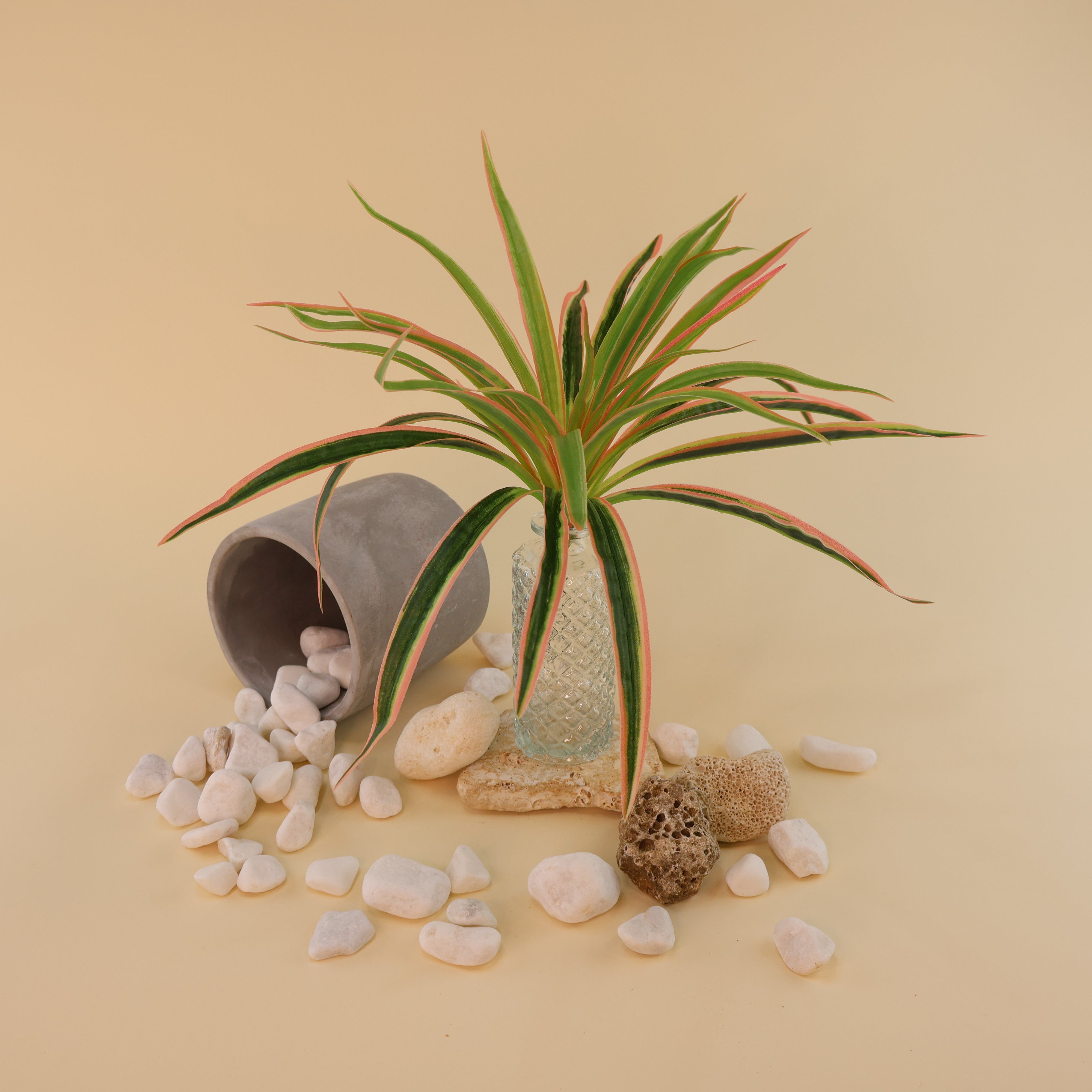 Künstliche Grünpflanze mit langen schmalen Blättern in Grün mit hellen und rosafarbenen Akzenten in einer Glasvase, dekorativ arrangiert mit weißen Dekosteinen und umgestürztem Blumentopf auf hellem Hintergrund.