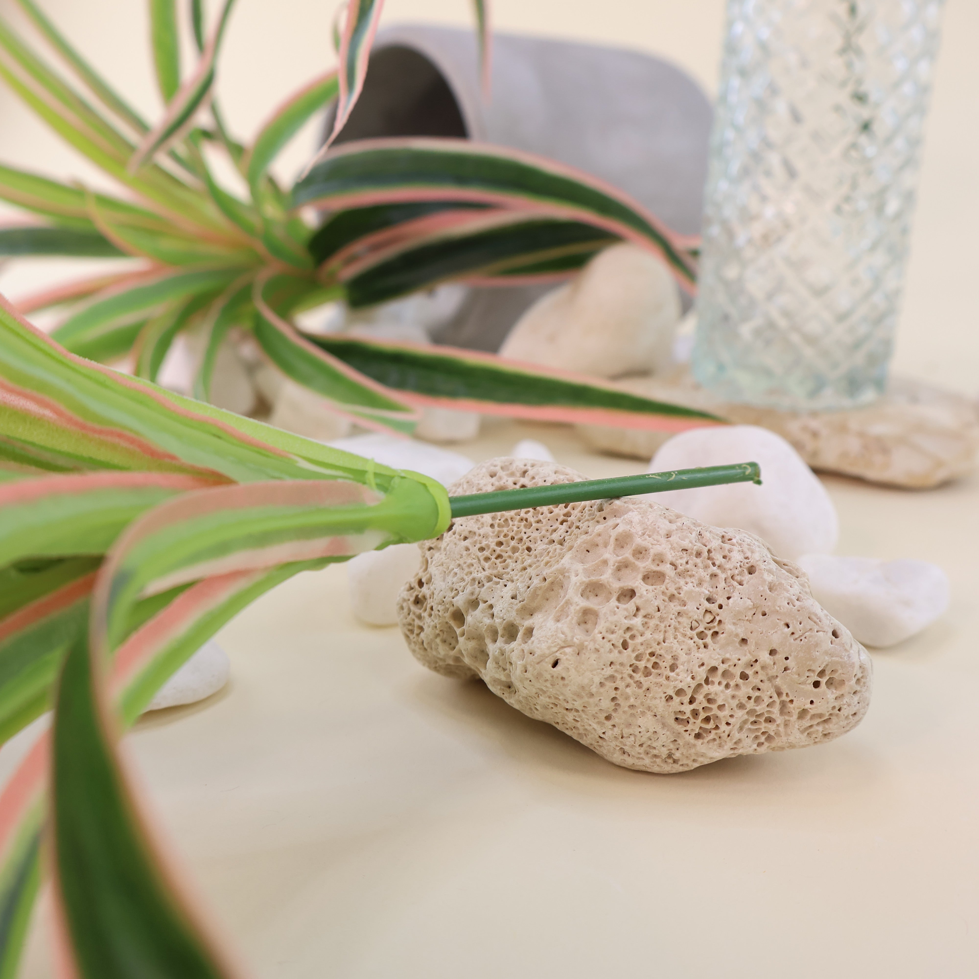 Detailaufnahme künstlicher Grünpflanzen mit langen schmalen Blättern in Grün mit hellen und rosafarbenen Akzenten neben Dekosteinen, Glasvase und umgestürztem Blumentopf auf hellem Hintergrund.