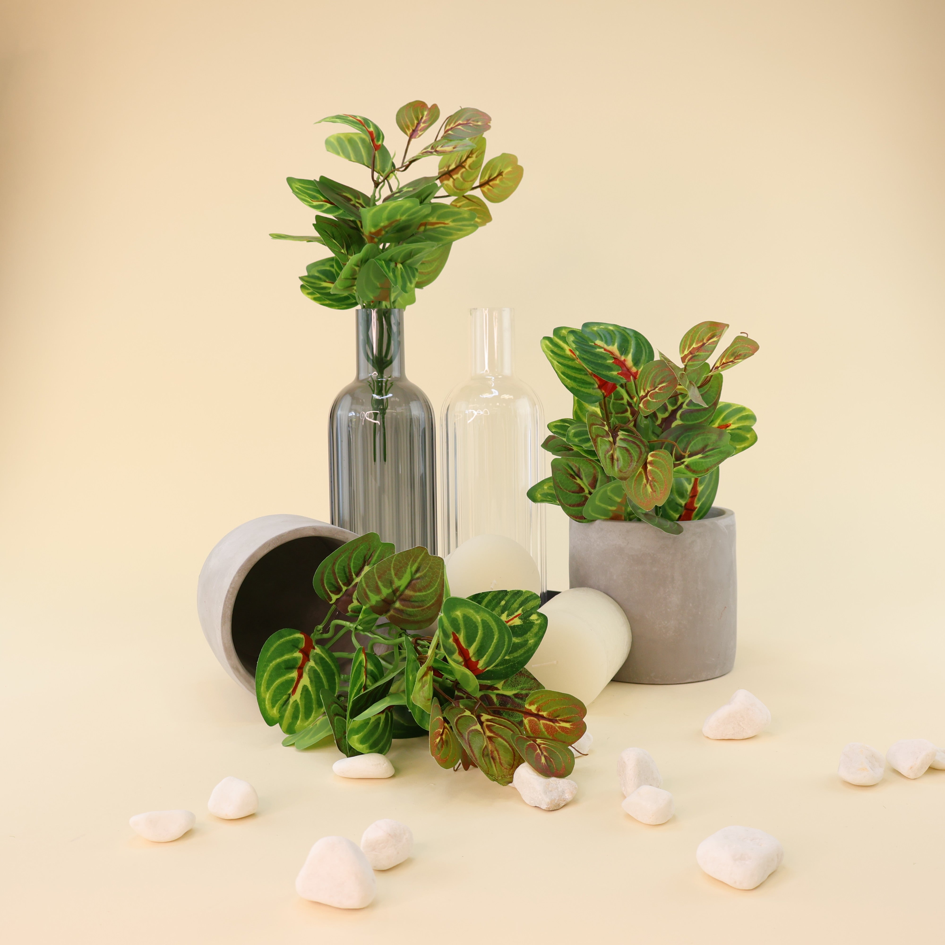 Künstliche Grünpflanzen mit dekorativen Blättern in verschiedenen Grün-, Oliv- und Rottönen stilvoll arrangiert mit Glasvasen, grauem Blumentopf, liegendem Keramiktopf, weißen Kerzen und Natursteinen. Die natürliche Blattzeichnung und die vielseitigen Dekorationsmöglichkeiten der Kunstpflanzen stehen im Mittelpunkt der harmonischen Wohnraumdekoration.