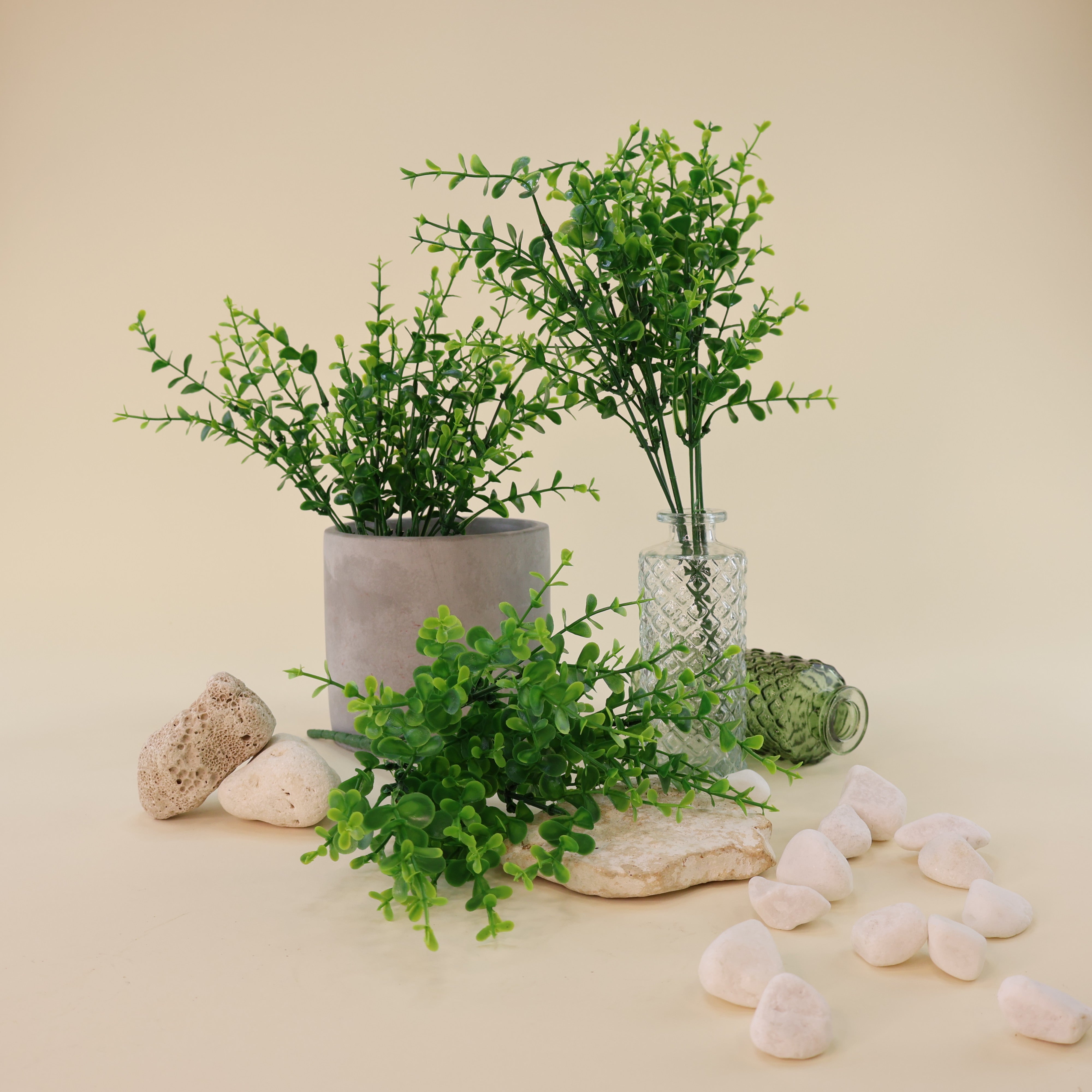 Mehrere künstliche Grünzweige mit kleinen runden Blättern in verschiedenen Grüntönen dekorativ arrangiert in grauem Blumentopf und strukturierter Glasvase, kombiniert mit weißen Dekosteinen und Natursteinen auf hellem Hintergrund.