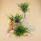 Mehrere künstliche Grünzweige mit kleinen runden Blättern in verschiedenen Grüntönen dekorativ arrangiert in grauem Blumentopf und großer Glasvase, kombiniert mit weißen Dekosteinen, Natursteinen und goldener Buddhafigur auf hellem Hintergrund.