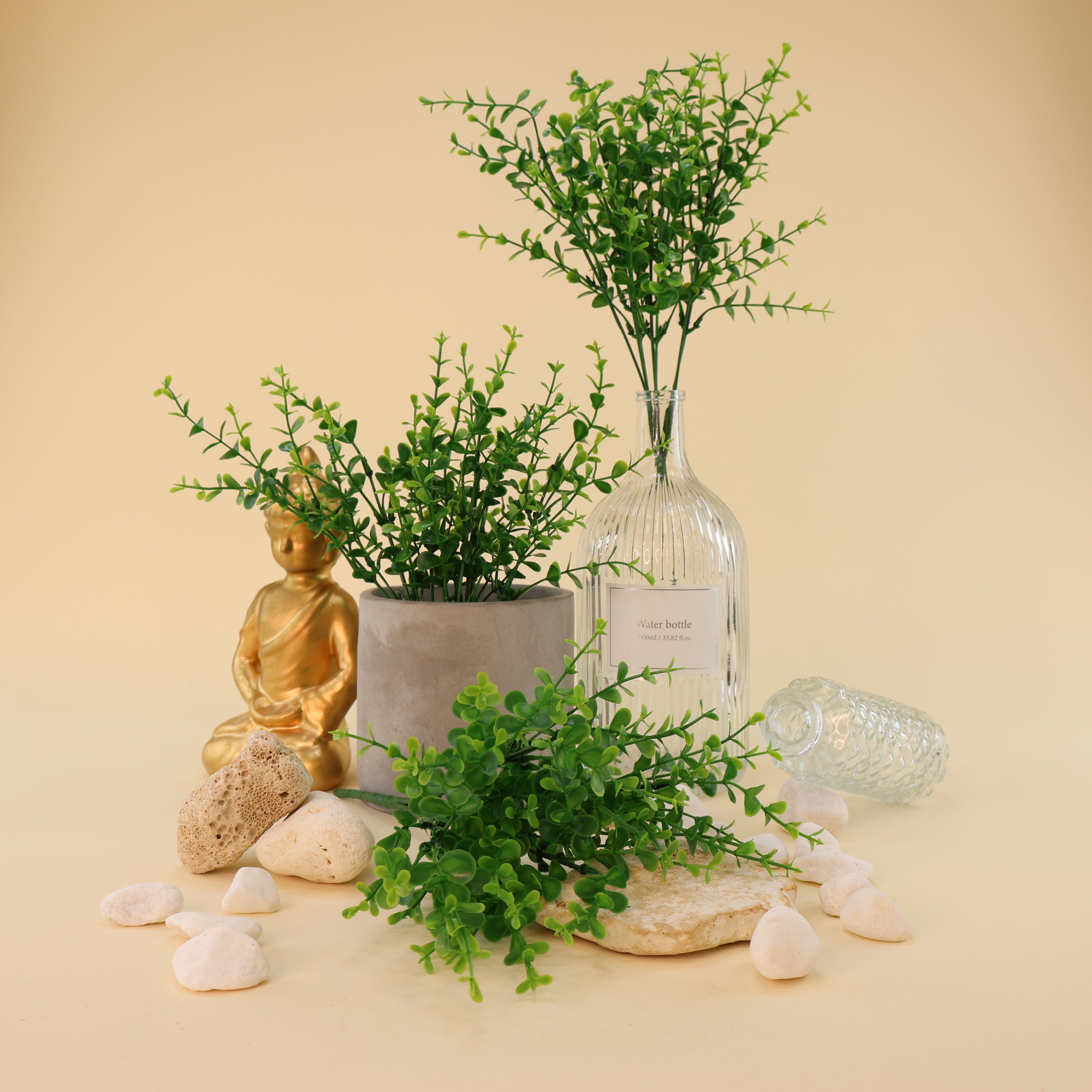 Mehrere künstliche Grünzweige mit kleinen runden Blättern in verschiedenen Grüntönen dekorativ arrangiert in grauem Blumentopf und großer Glasvase, kombiniert mit weißen Dekosteinen, Natursteinen und goldener Buddhafigur auf hellem Hintergrund.