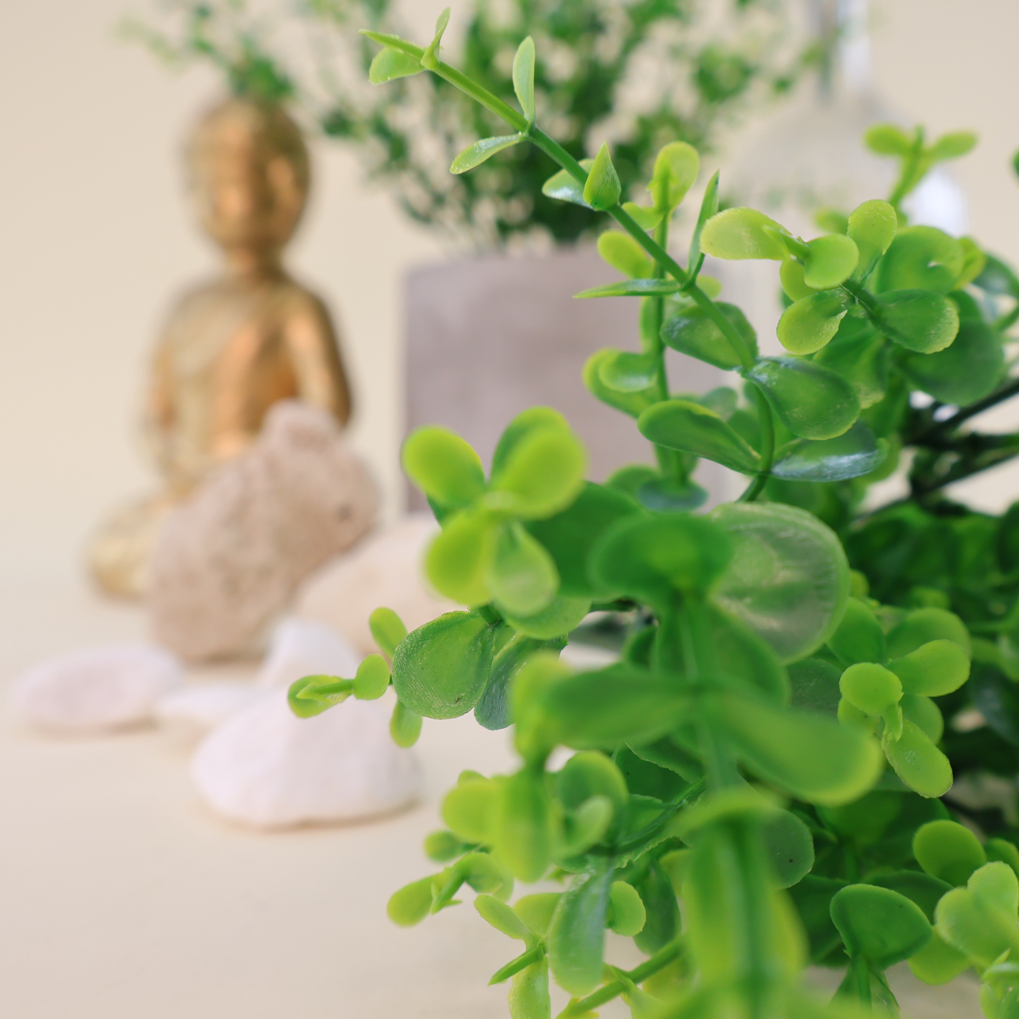 Makroaufnahme künstlicher Grünzweige mit kleinen runden Blättern in verschiedenen Grüntönen, dekorativ arrangiert mit Natursteinen, grauem Blumentopf und goldener Buddhafigur im Hintergrund.