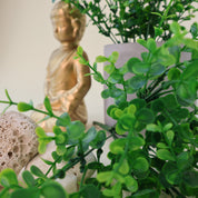 Dekorative Arrangementszene mit künstlichen Grünzweigen in verschiedenen Grüntönen, kombiniert mit grauem Blumentopf, Natursteinen und goldener Buddhafigur auf hellem Hintergrund.