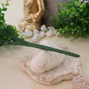 Detailaufnahme dekorativer Arrangementszene mit künstlichen Grünzweigen, weißen Natursteinen, grauem Blumentopf und goldener Buddhafigur auf hellem Hintergrund.