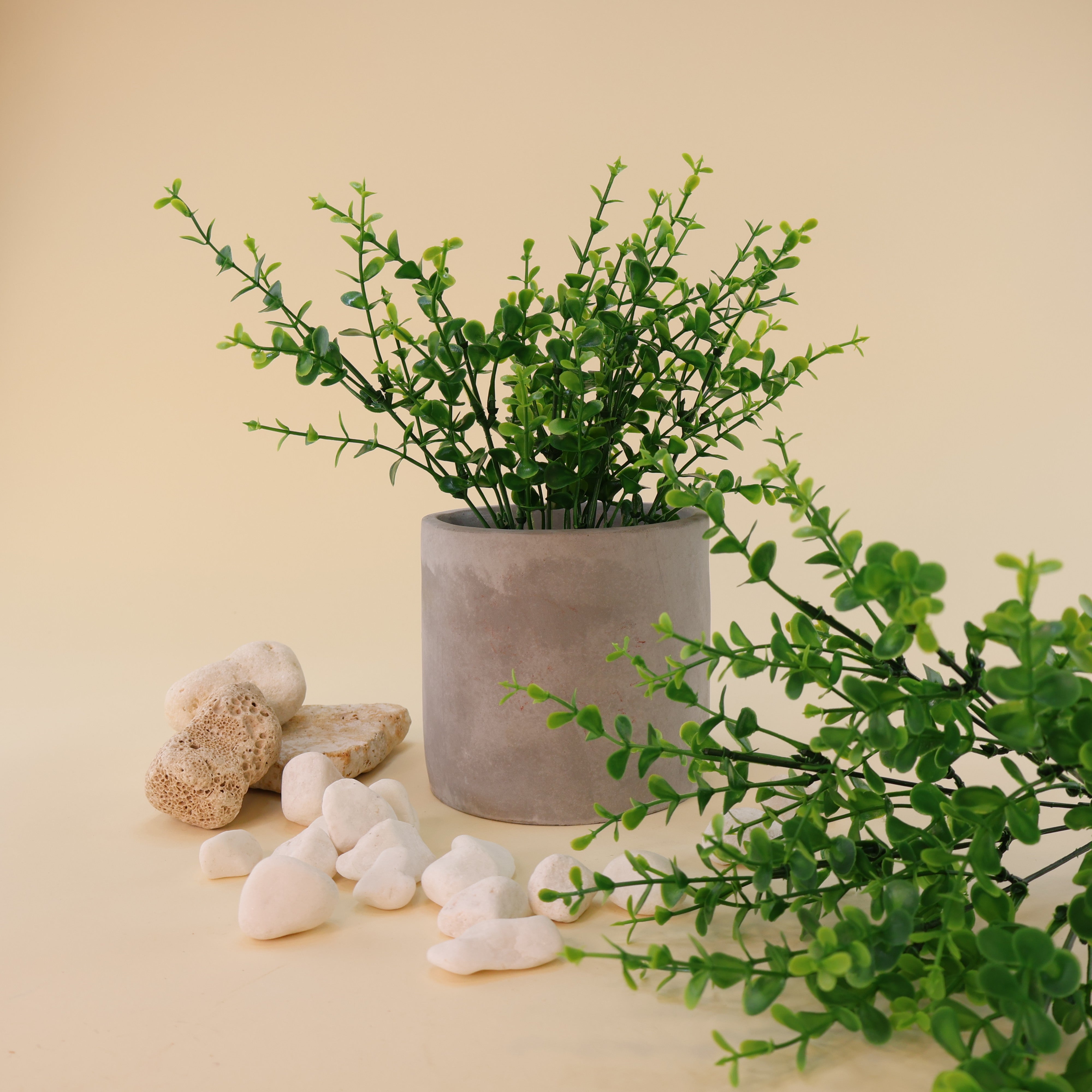 Künstliche Grünzweige mit kleinen runden Blättern in verschiedenen Grüntönen dekorativ arrangiert in einem grauen Blumentopf, kombiniert mit weißen Dekosteinen und Natursteinen auf hellem Hintergrund.