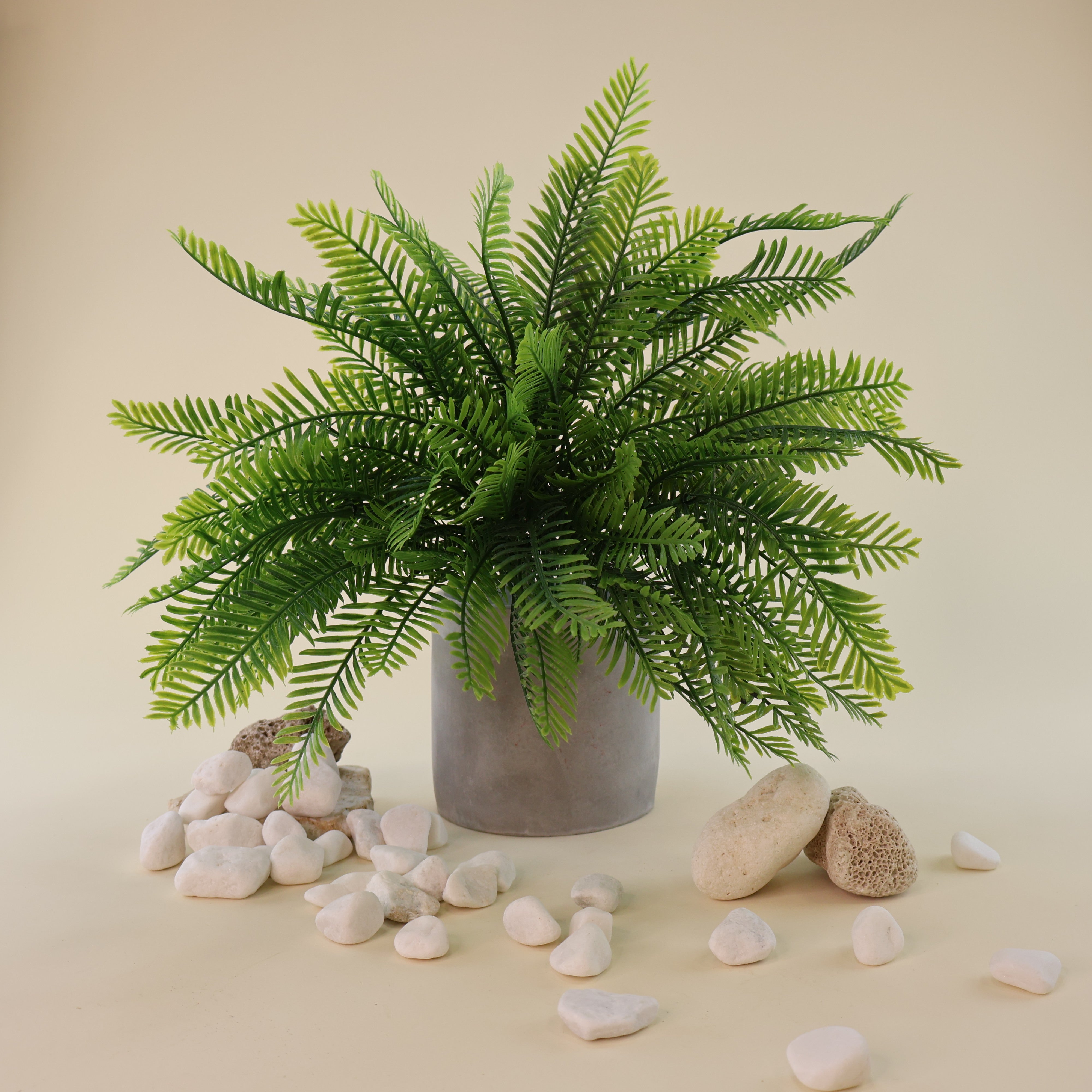 Künstlicher Farnbusch mit filigranen, naturgetreu gestalteten Farnwedeln in verschiedenen Grüntönen in einem schlichten grauen Blumentopf. Weiße Natursteine und helle Dekoelemente ergänzen das üppige Arrangement und unterstreichen die natürliche Ausstrahlung der Kunstpflanze.