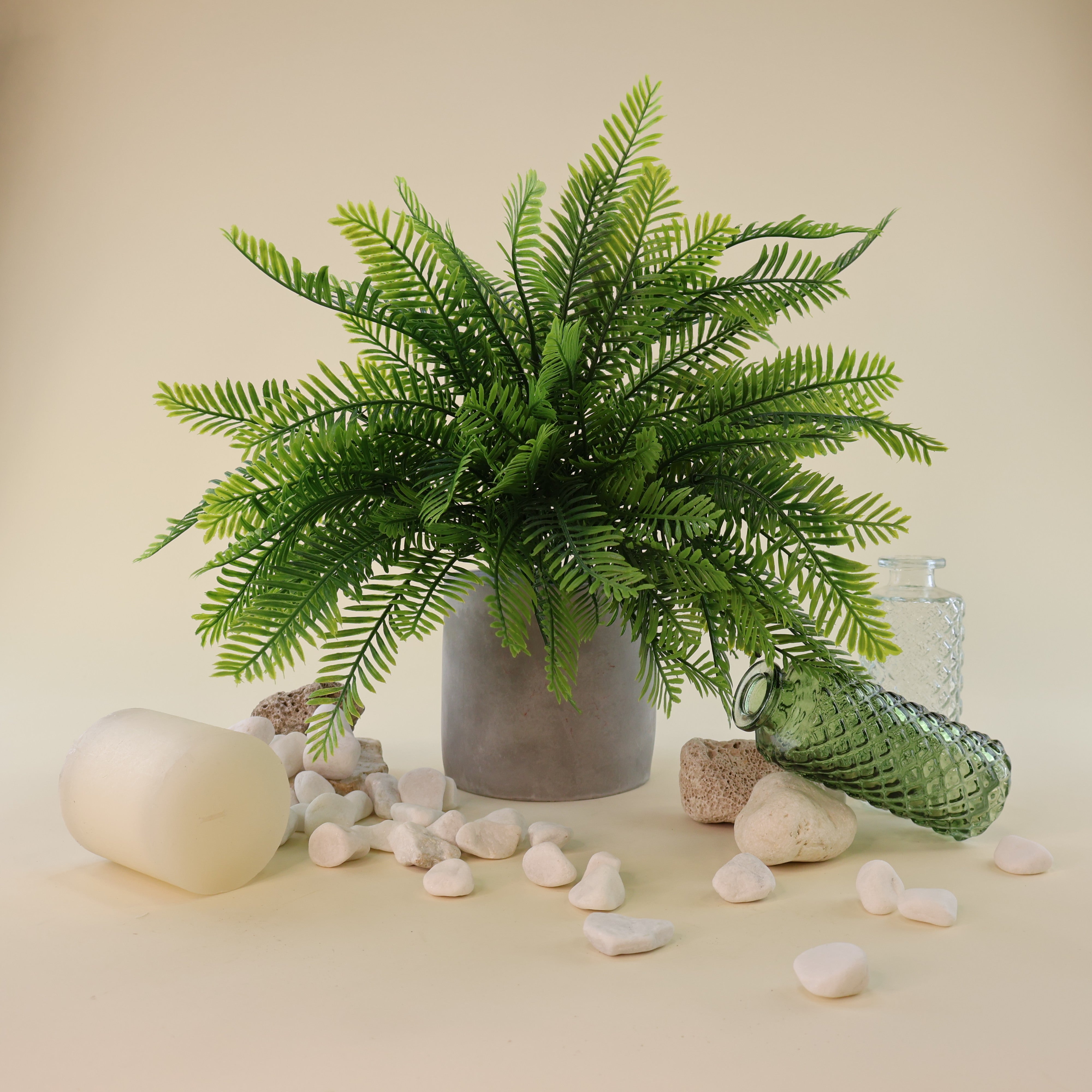 Künstlicher Farnbusch mit filigranen, naturgetreu gestalteten Farnwedeln in verschiedenen Grüntönen in einem grauen Blumentopf. Das üppige Arrangement wird von weißen Natursteinen, dekorativen Glasvasen und einer Kerze ergänzt und eignet sich ideal für moderne Wohn- und Geschäftsräume.