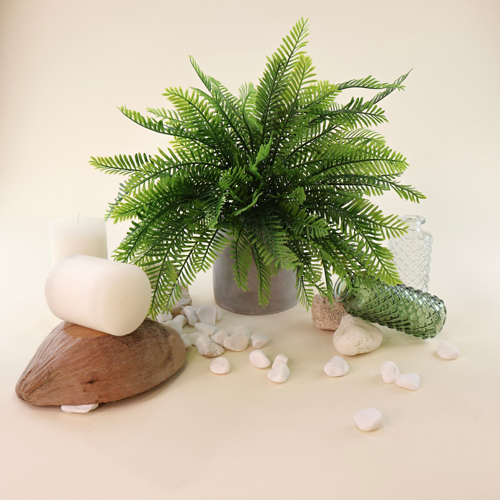 Üppiger künstlicher Farnbusch mit fein gefiederten Farnwedeln in verschiedenen Grüntönen in einem schlichten grauen Blumentopf. Das naturinspirierte Arrangement wird durch weiße Natursteine, strukturierte Glasvasen, Kerzen und ein dekoratives Holzstück ergänzt und sorgt für eine frische, moderne Wohnatmosphäre.
