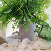 Künstlicher Farnbusch mit filigranen, naturgetreu gestalteten Farnwedeln in verschiedenen Grüntönen in einem schlichten grauen Blumentopf. Weiße Natursteine, eine grüne Glasvase und dezente Dekoelemente ergänzen das Arrangement und unterstreichen die natürliche Ausstrahlung der Kunstpflanze.