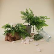 Künstliche Farnzweige mit filigranen, naturgetreu gestalteten Farnwedeln in verschiedenen Grüntönen als dekoratives Arrangement. Die Kunstpflanzen sind in einem grauen Blumentopf und auf einem rustikalen Holzstück arrangiert, ergänzt durch Natursteine, Glasvasen und eine Kerze für eine natürliche Wohnraumdekoration.
