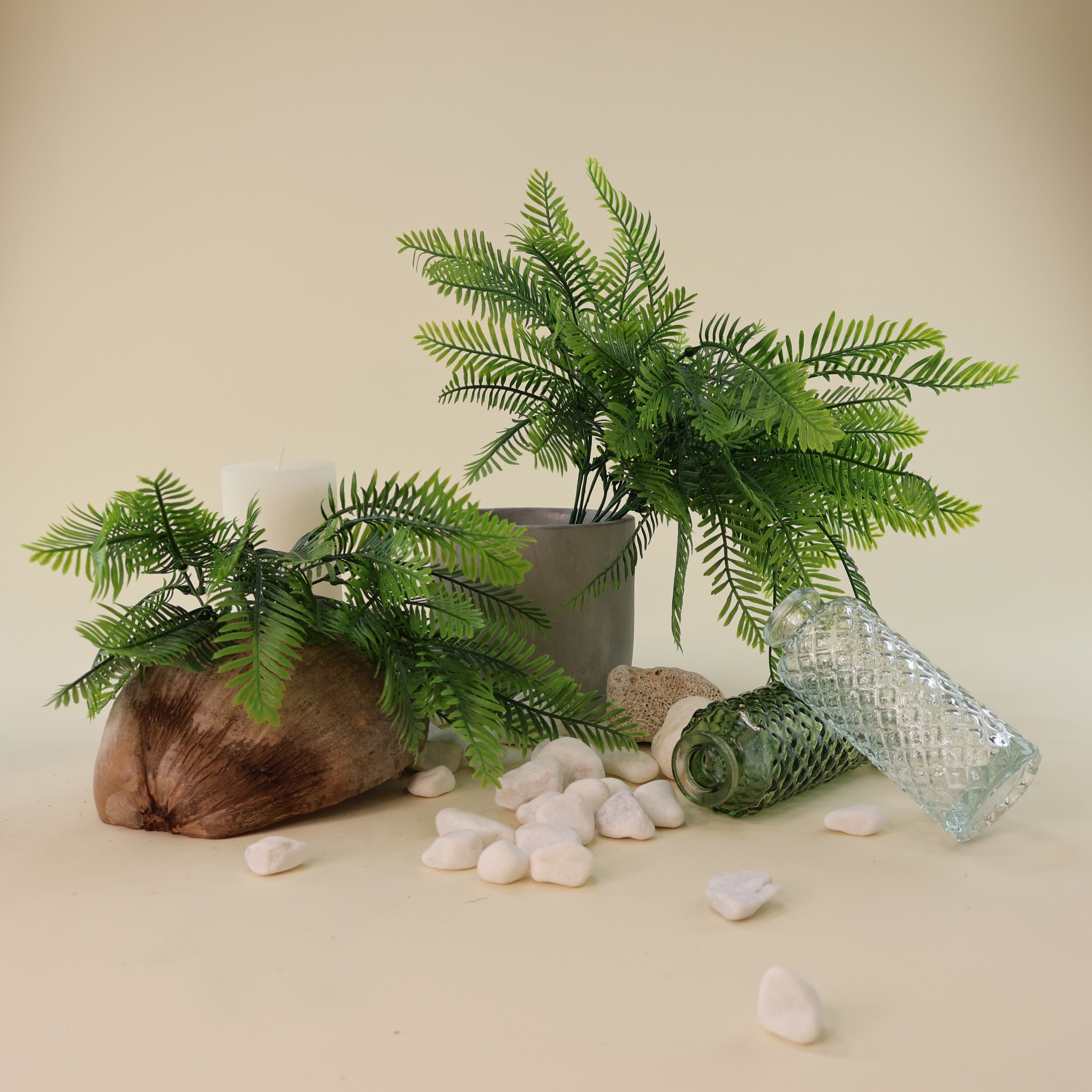 Künstliche Farnzweige mit filigranen, naturgetreu gestalteten Farnwedeln in verschiedenen Grüntönen als dekoratives Arrangement. Die Kunstpflanzen sind in einem grauen Blumentopf und auf einem rustikalen Holzstück arrangiert, ergänzt durch Natursteine, Glasvasen und eine Kerze für eine natürliche Wohnraumdekoration.