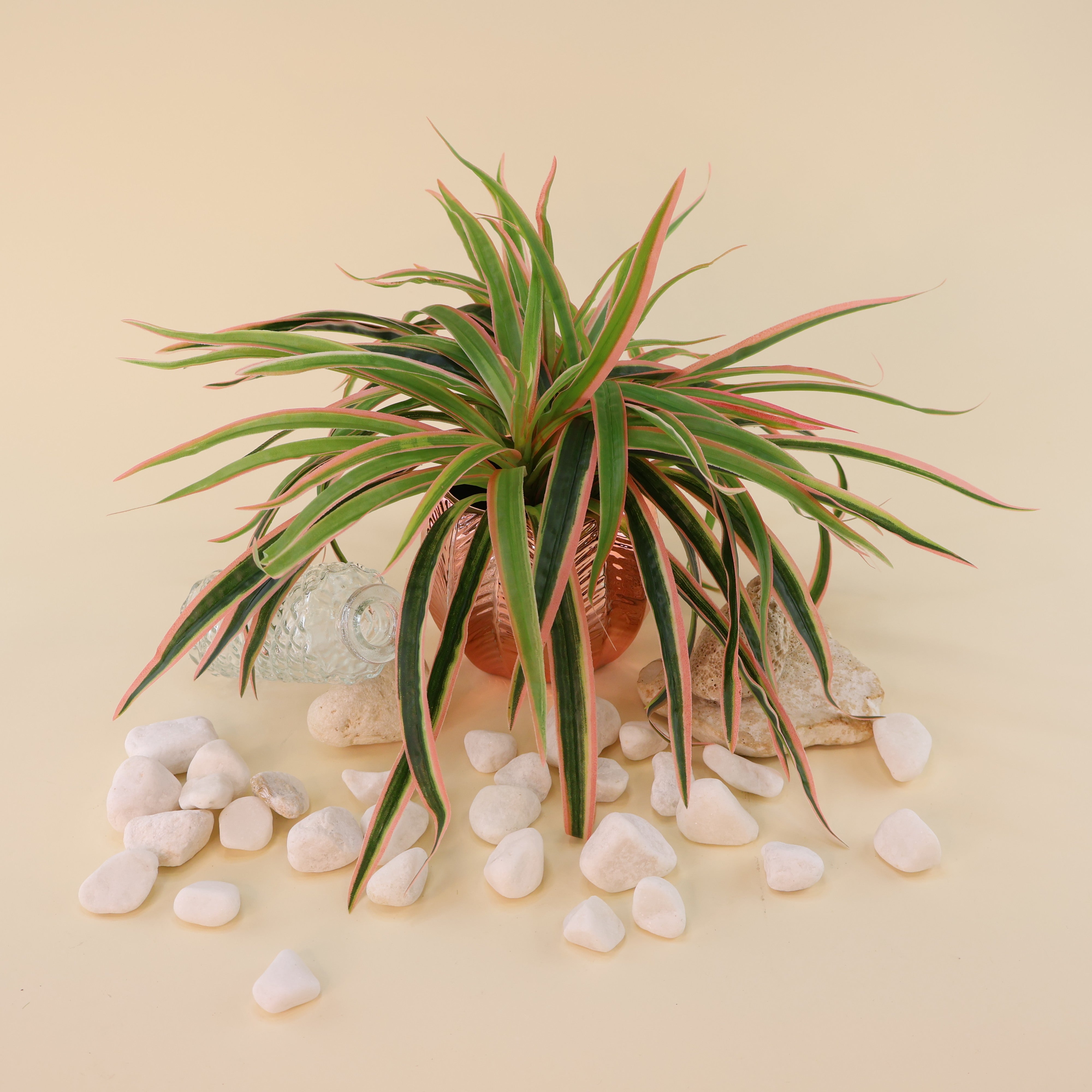 Künstliche Grünpflanze mit langen schmalen Blättern in Grün mit hellen und rosafarbenen Akzenten in einem dekorativen Blumentopf, arrangiert mit weißen Dekosteinen und Glasvase auf hellem Hintergrund.
