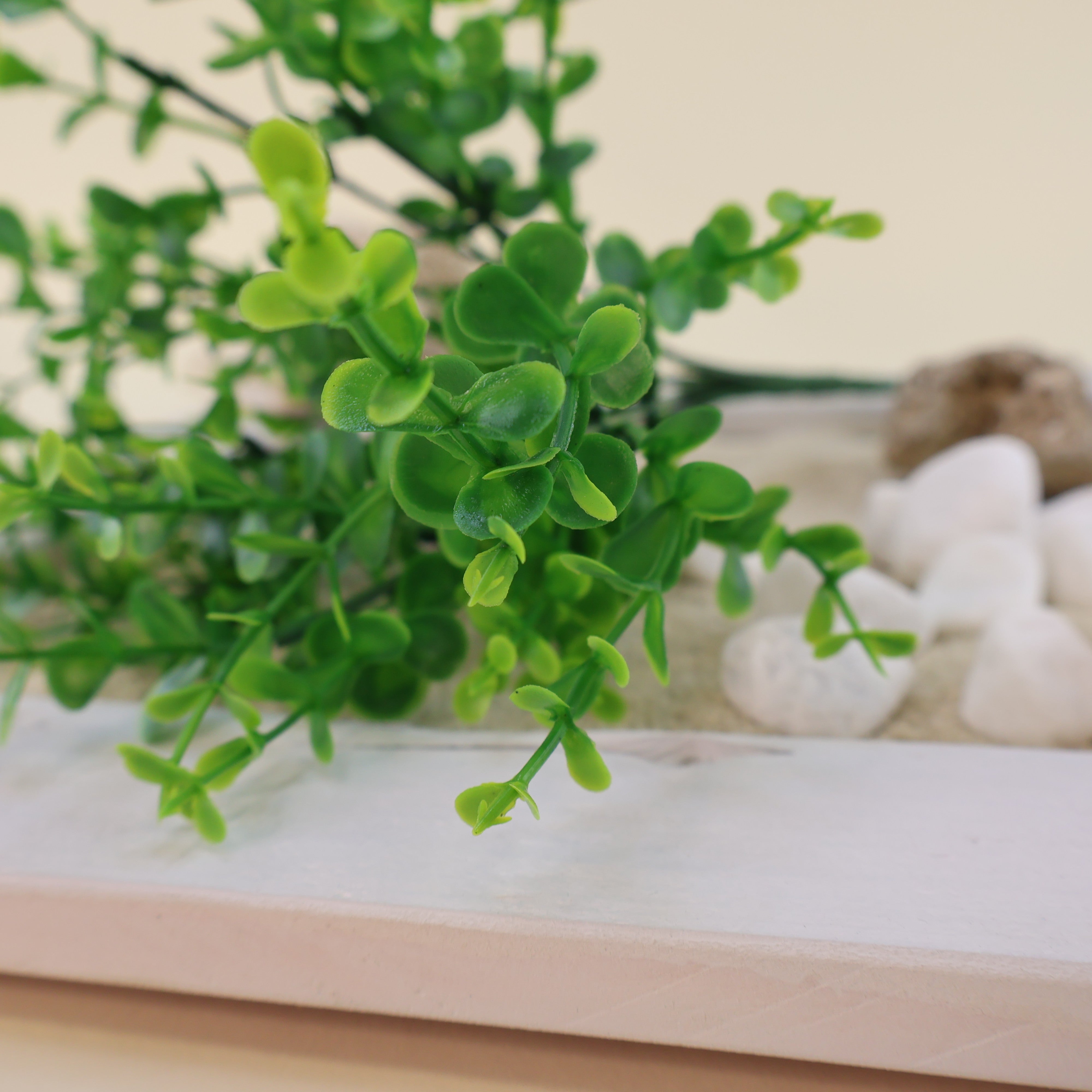 Detailaufnahme künstlicher Grünzweige mit kleinen runden Blättern in verschiedenen Grüntönen, dekorativ arrangiert mit weißen Dekosteinen und hellem Sand auf weißem Holztablett.