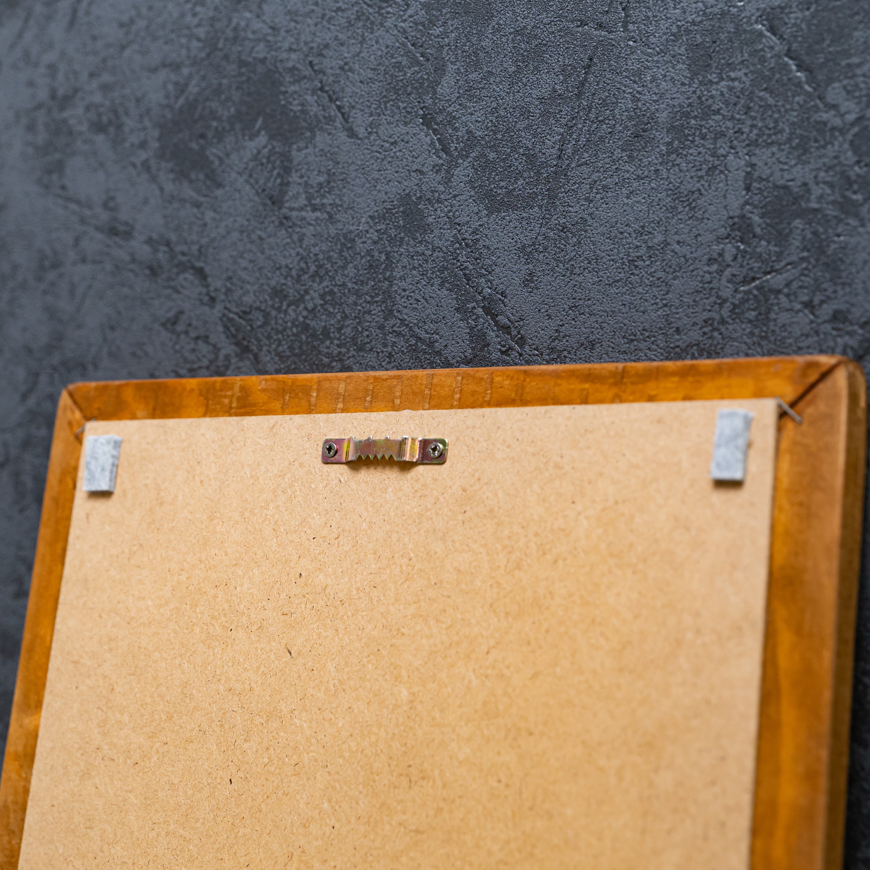 Rückseite eines Holzrahmens mit MDF-Platte und mittig montierter Zackenaufhängung vor einer dunklen Strukturwand