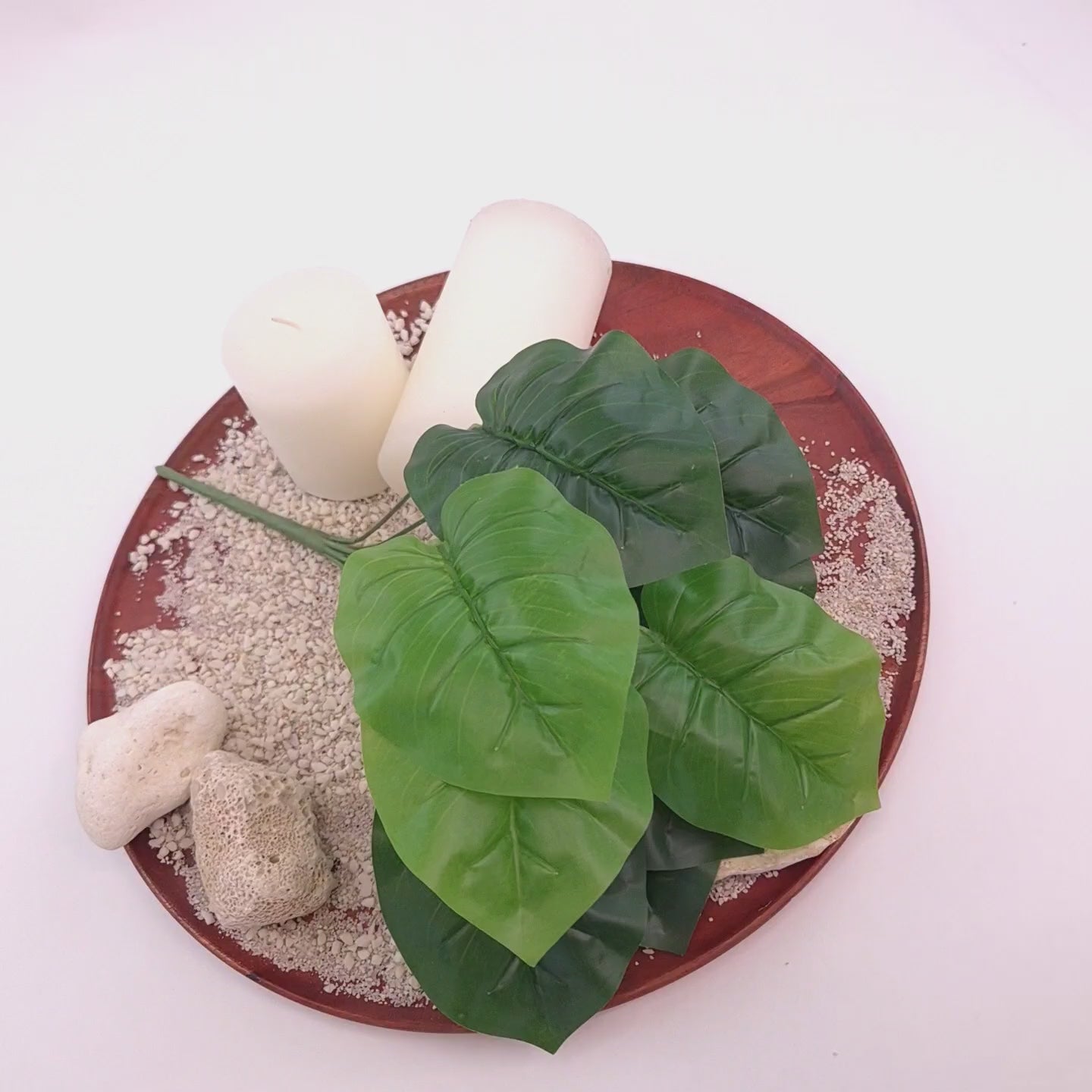 Kurzes Produktvideo einer künstlichen Grünpflanze mit großen Blättern in Hell- und Dunkelgrün auf einem dekorativen Tablett mit Sand, Natursteinen und weißen Kerzen. Die Kamera fährt langsam von der Gesamtansicht zu detailreichen Nahaufnahmen der Blätter und Oberflächenstruktur.